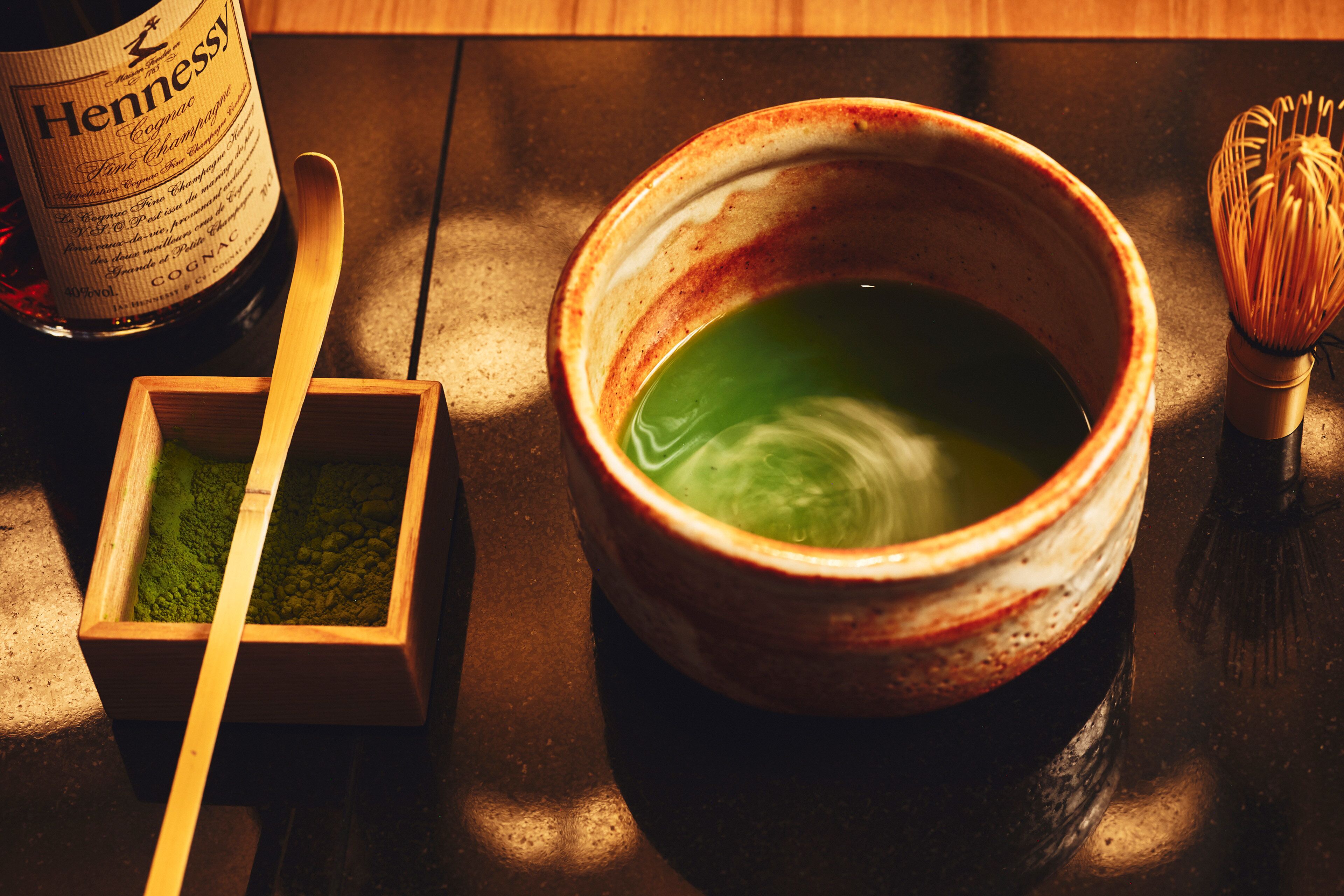 A wooden box with green powder, a ceramic bowl with green liquid, and a whisk on a wooden surface.