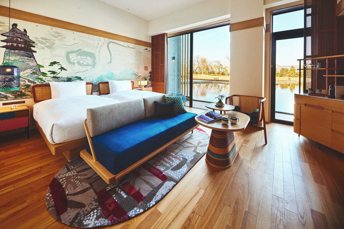 A modern hotel room with a large bed, a blue sofa, a small table, and a view of a lake through a sliding door.