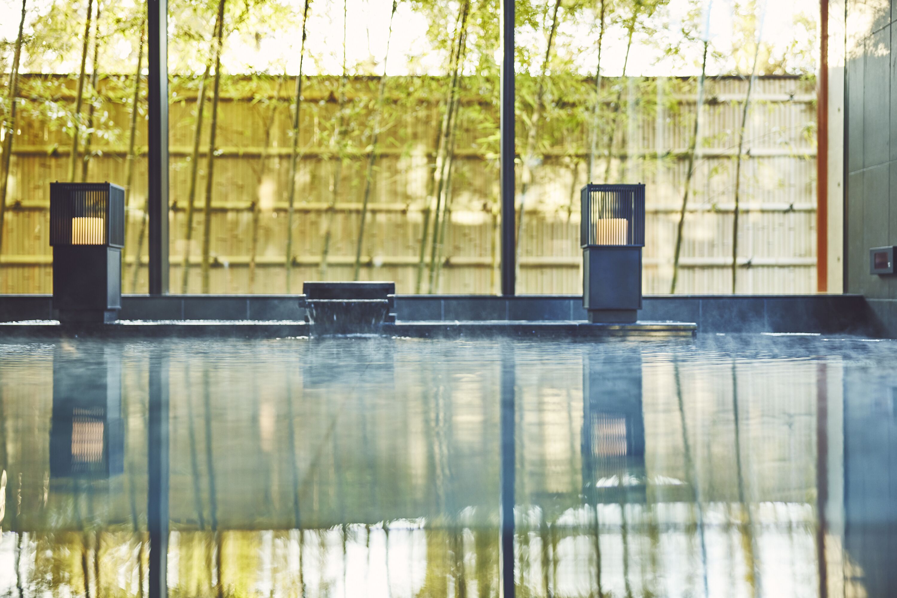 A serene indoor pool area with a bench and two wall-mounted lights, surrounded by a bamboo fence and large windows.