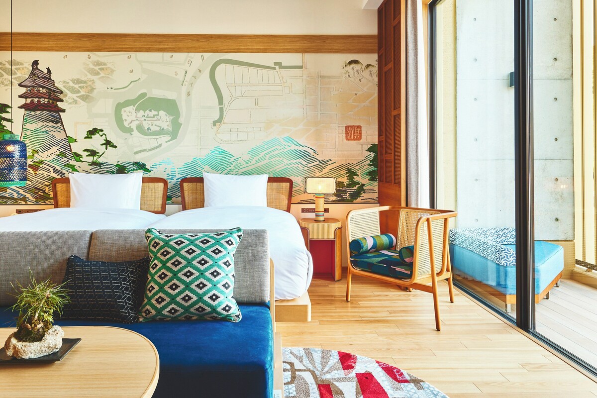 A hotel room with a large bed, a sofa, a chair, a small table with a lamp, and a large window.