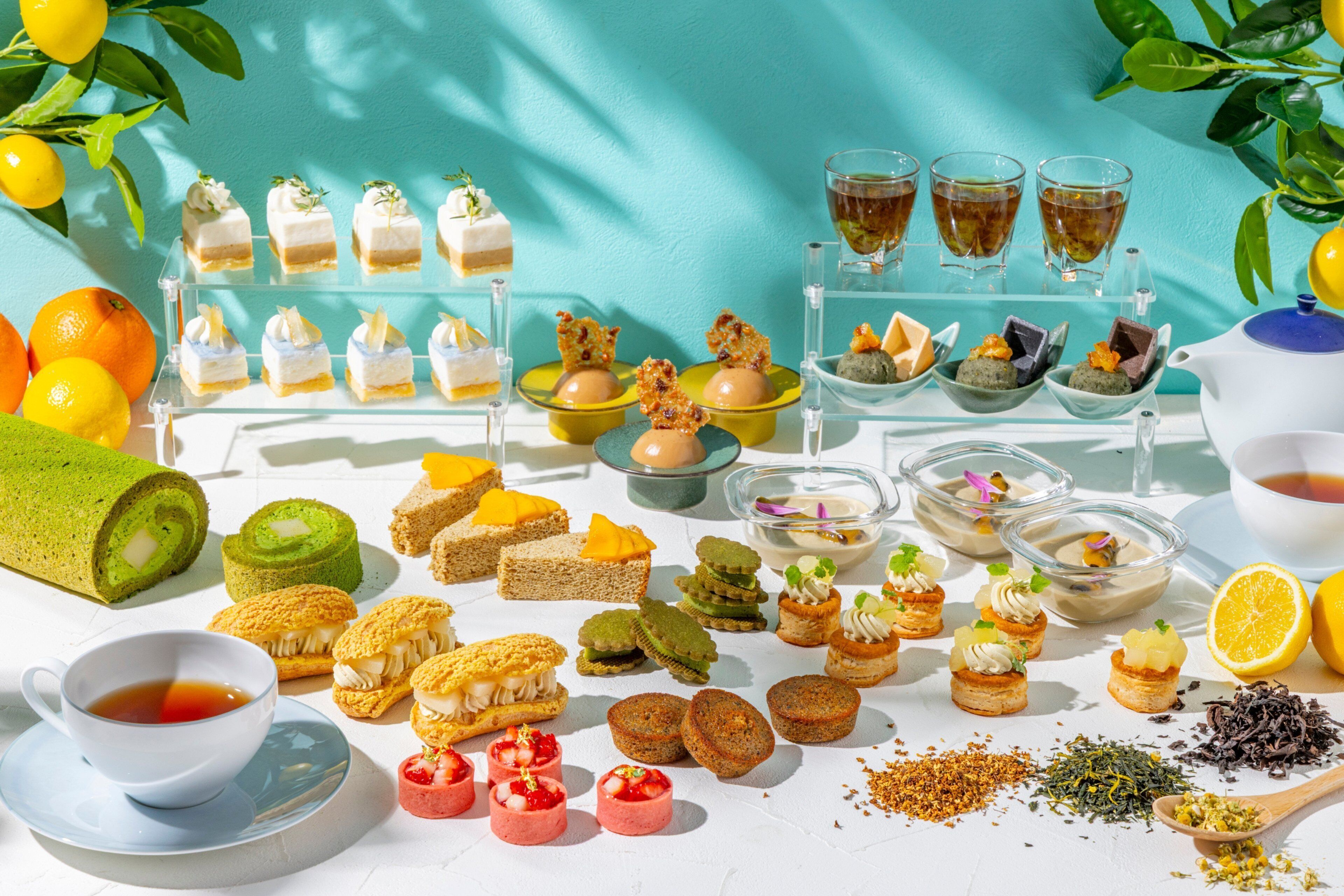 A variety of desserts and beverages on a table with a clear glass display case and a white teapot.