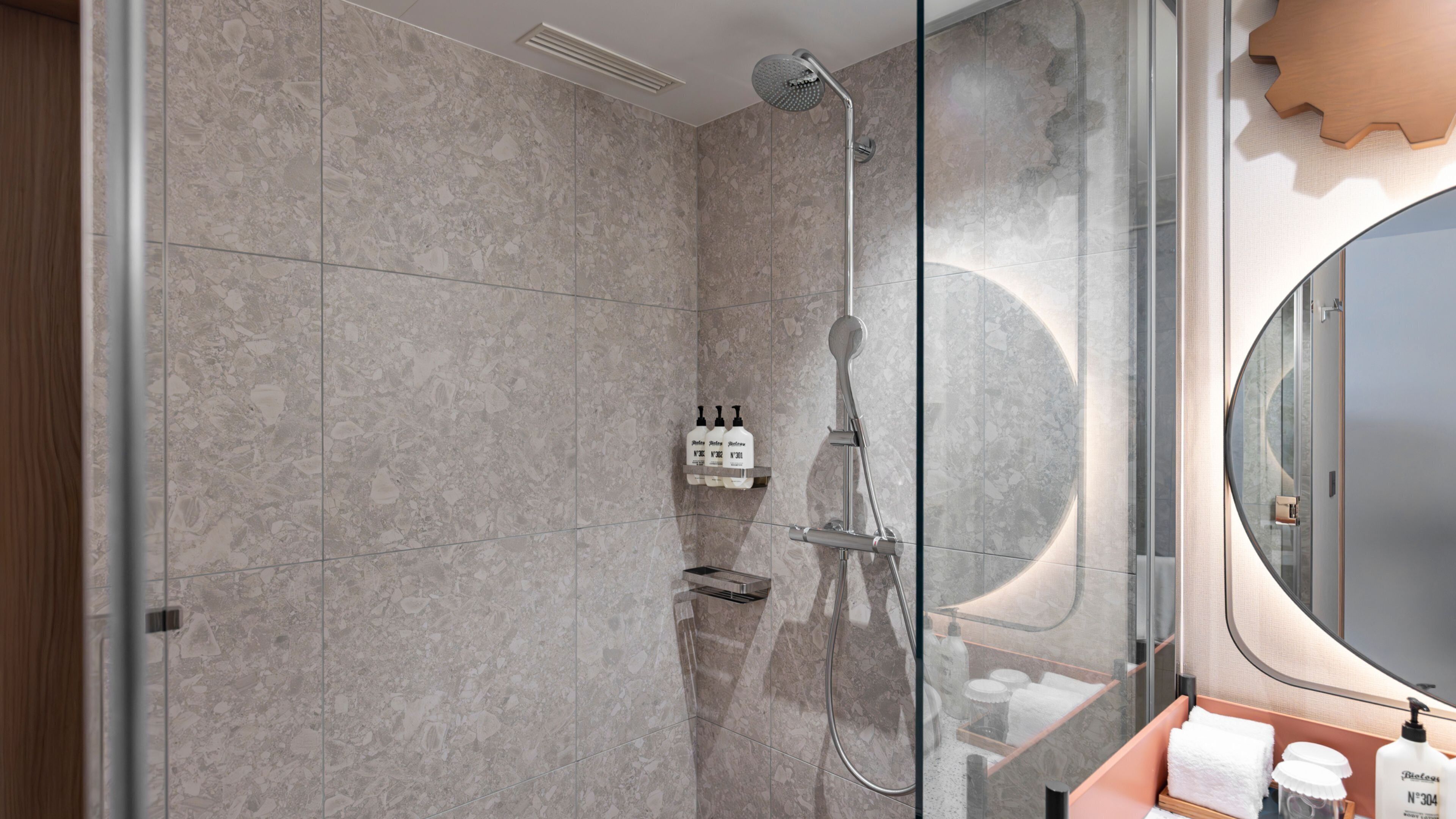 A modern bathroom with a glass shower enclosure, marble tiles, a vanity with a mirror, and toiletries.