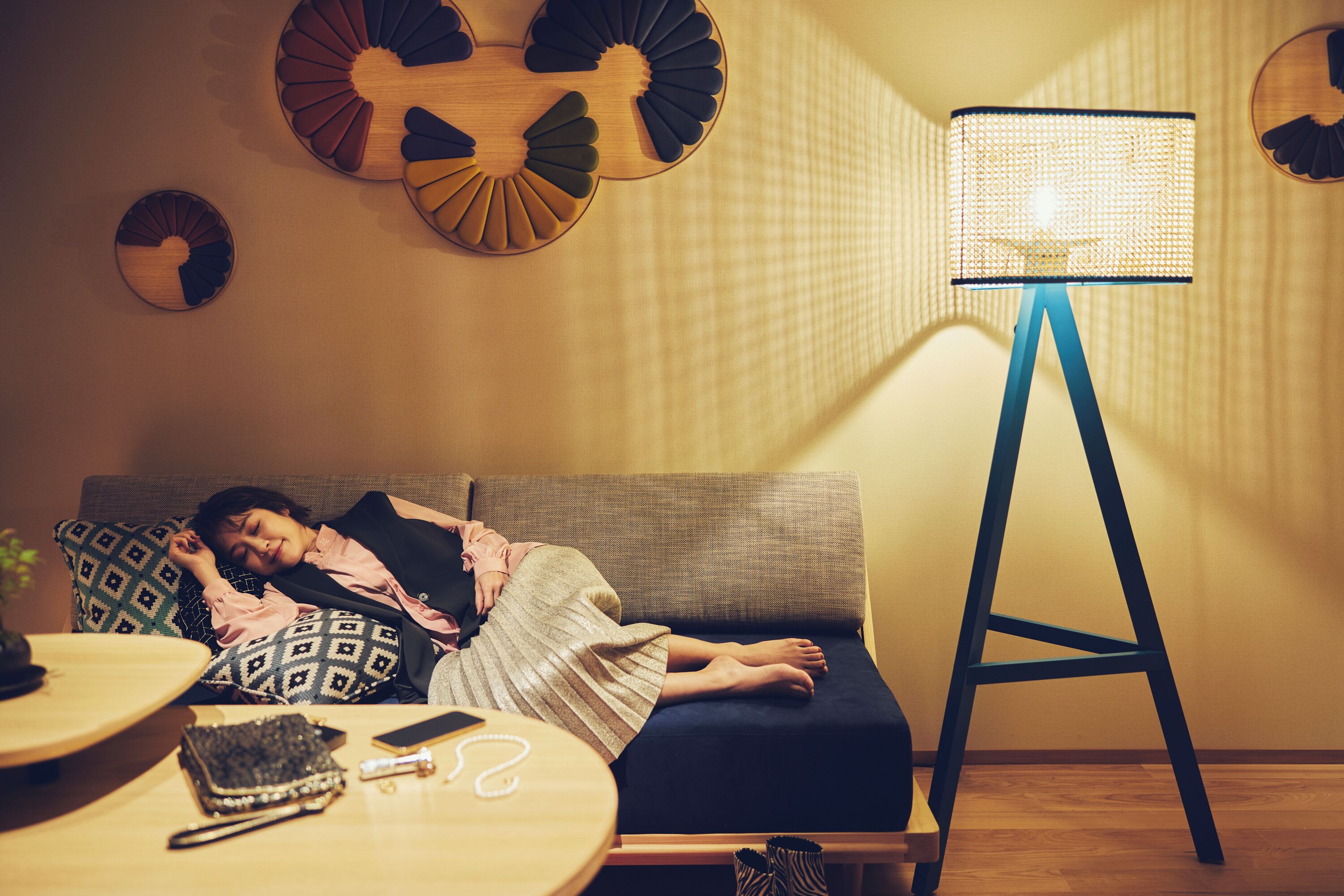 Une personne est endormie sur un canapé dans une pièce où se trouvent un lampadaire, un coussin à motifs et une petite table sur laquelle reposent un téléphone et un portefeuille.