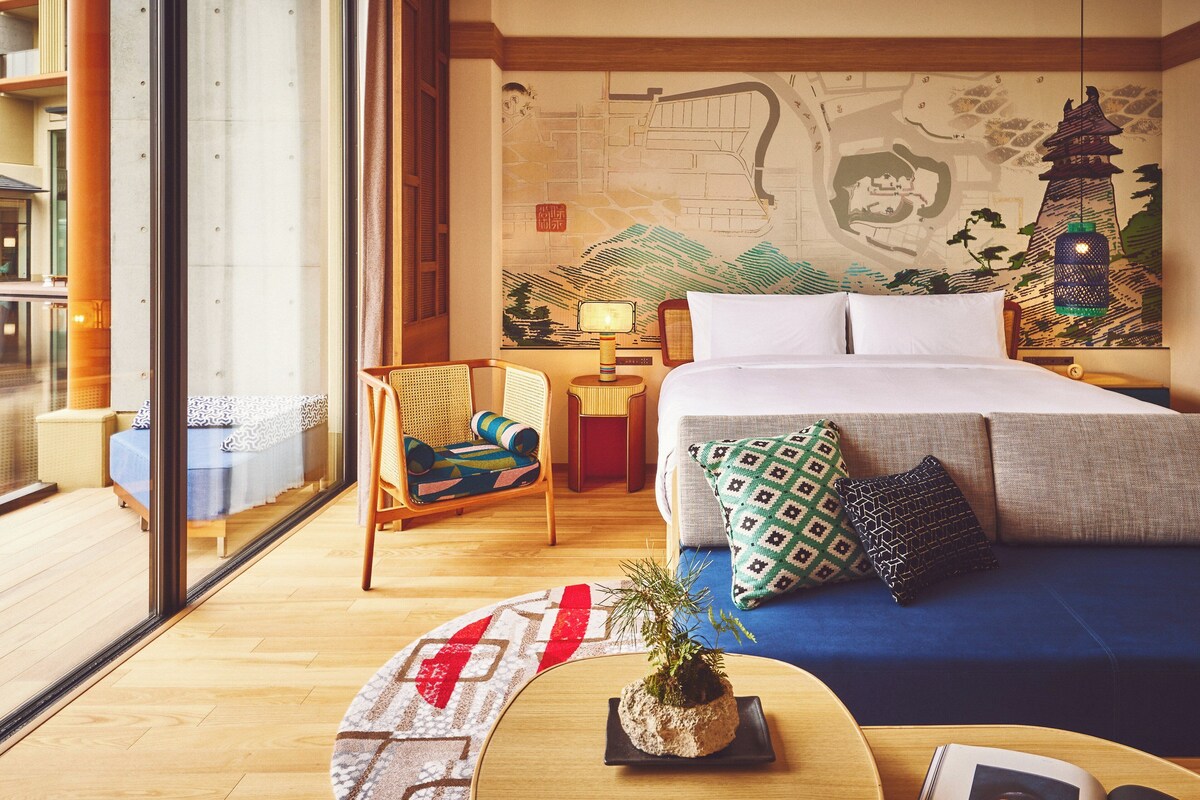 A modern hotel room with a large bed, a seating area with a chair and a small table, a large window, and a decorative wall mural.