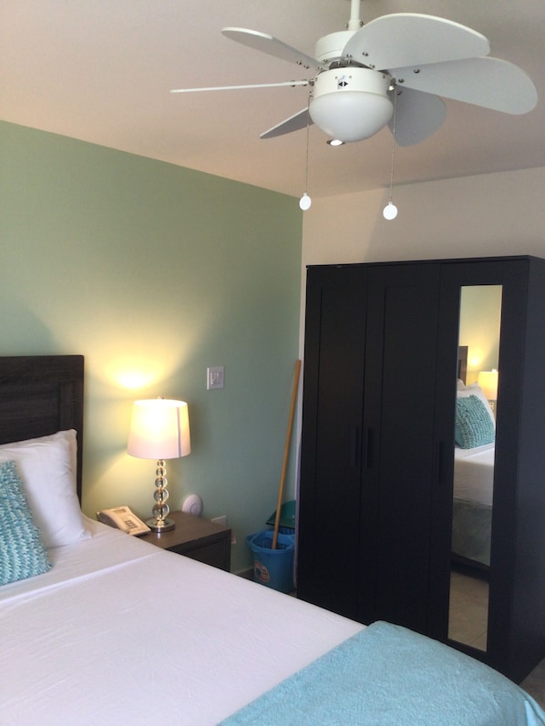 Quarto de hotel com cama, mesa de cabeceira com abajur, ventoinha no teto e guarda-roupa.