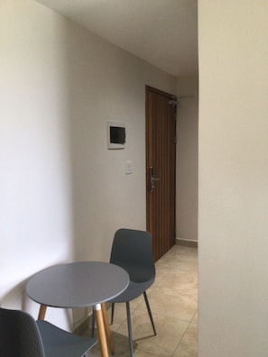 Um quarto pequeno e estreito, com uma mesa redonda e duas cadeiras, uma porta de madeira e um pequeno quadro na parede.