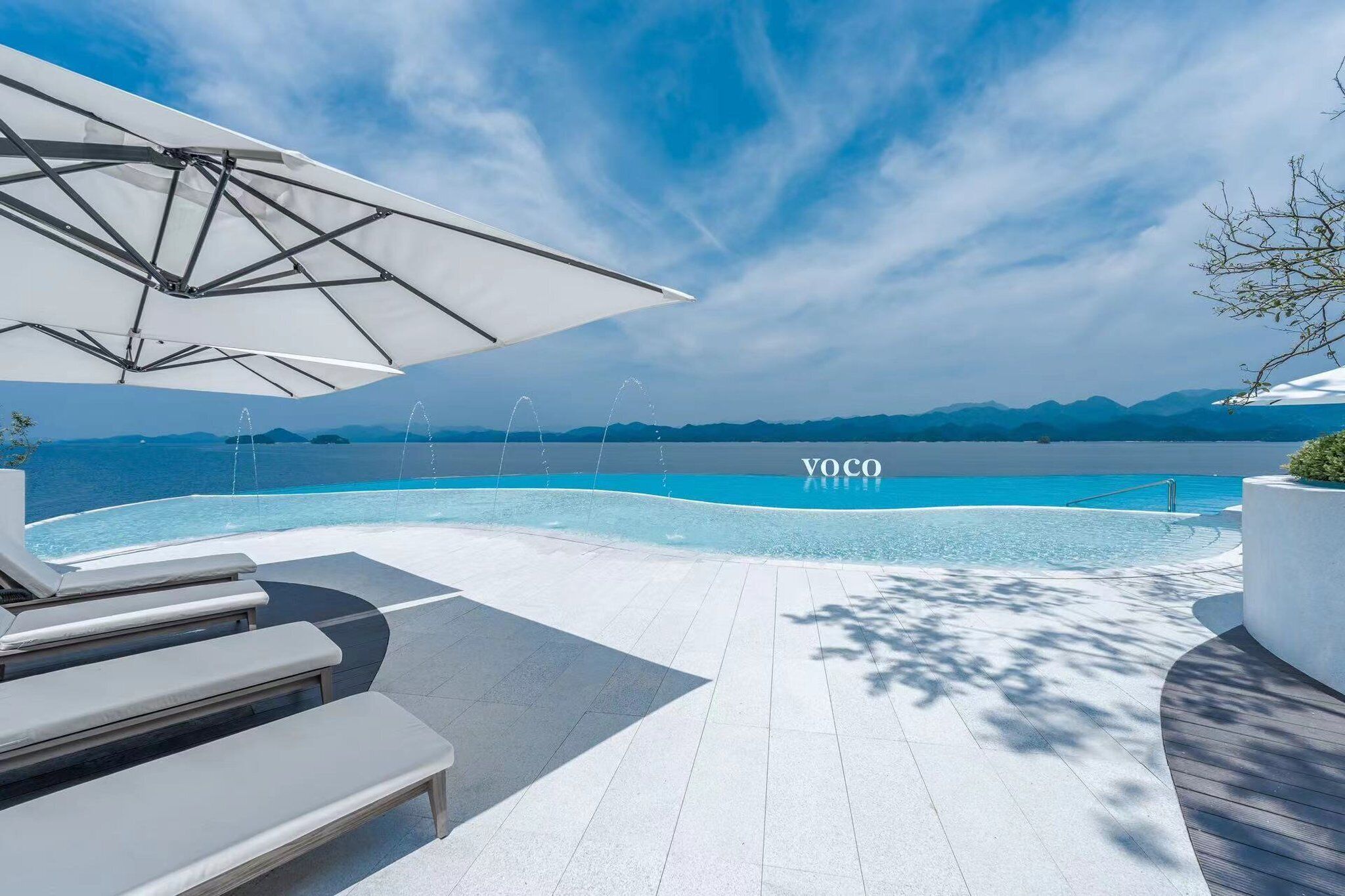 Een modern buitenzwembad met ligbedden en parasols, met uitzicht op een helderblauw meer en bergen.