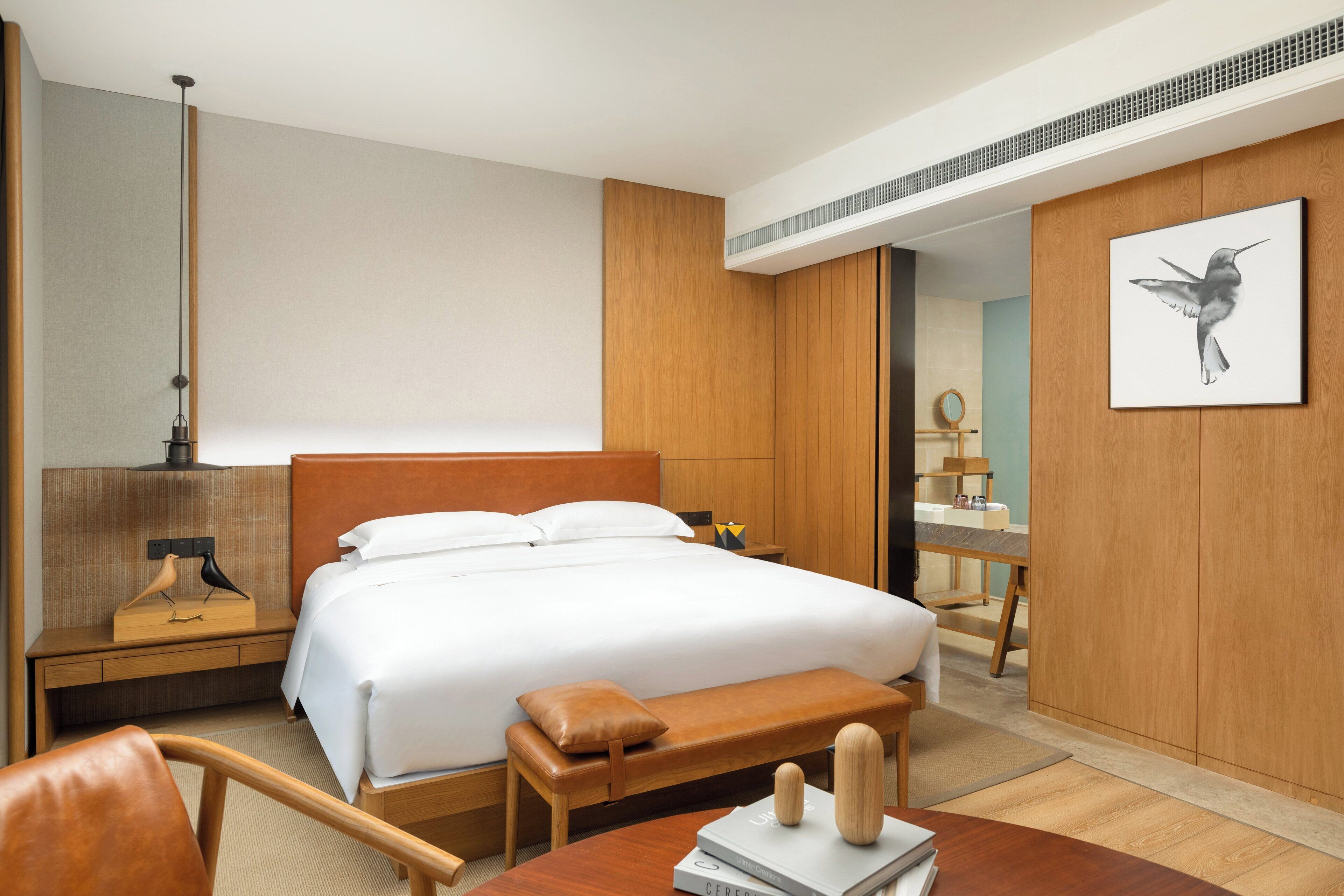 Une chambre d’hôtel avec un grand lit, une tête de lit en bois, un banc en bois, une table de chevet avec une lampe, un miroir et une œuvre d’art encadrée accrochée au mur.