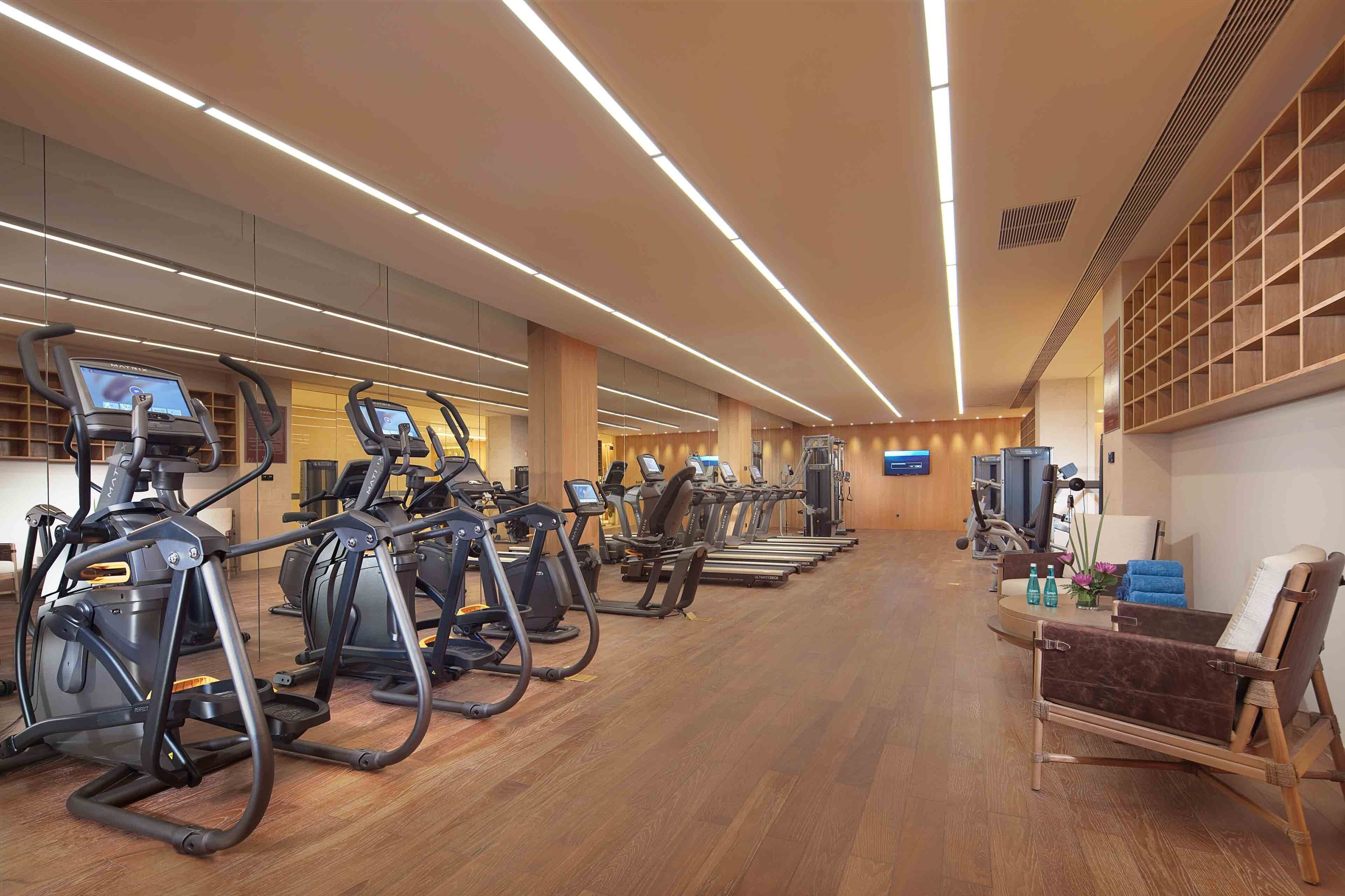 Une salle de sport équipée de plusieurs tapis de course et de plusieurs appareils elliptiques, dotée d’un plancher en bois et d’un espace détente avec un banc et une table.