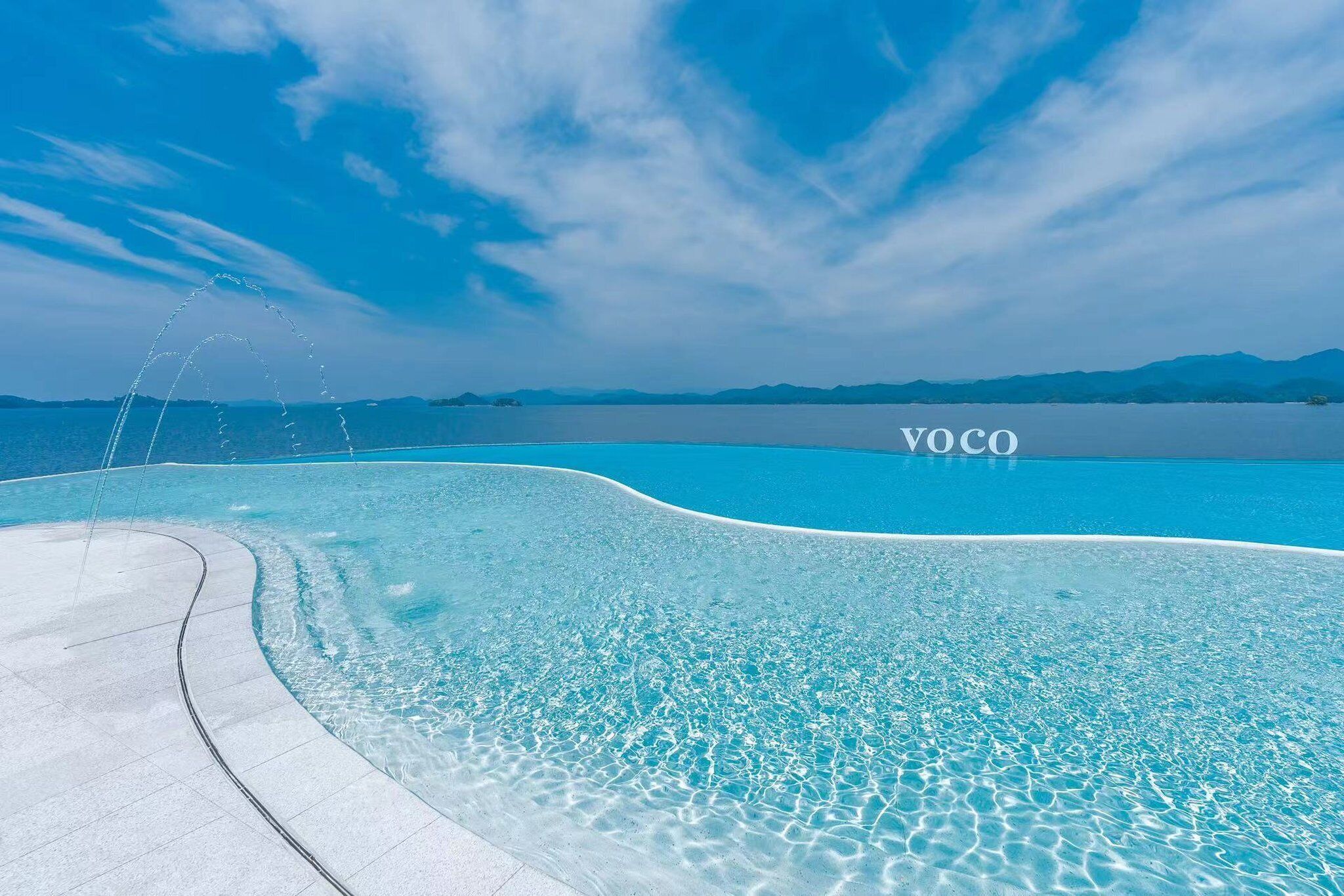 Una piscina azul cristalina con borde curvo, rodeada de una zona pavimentada.
