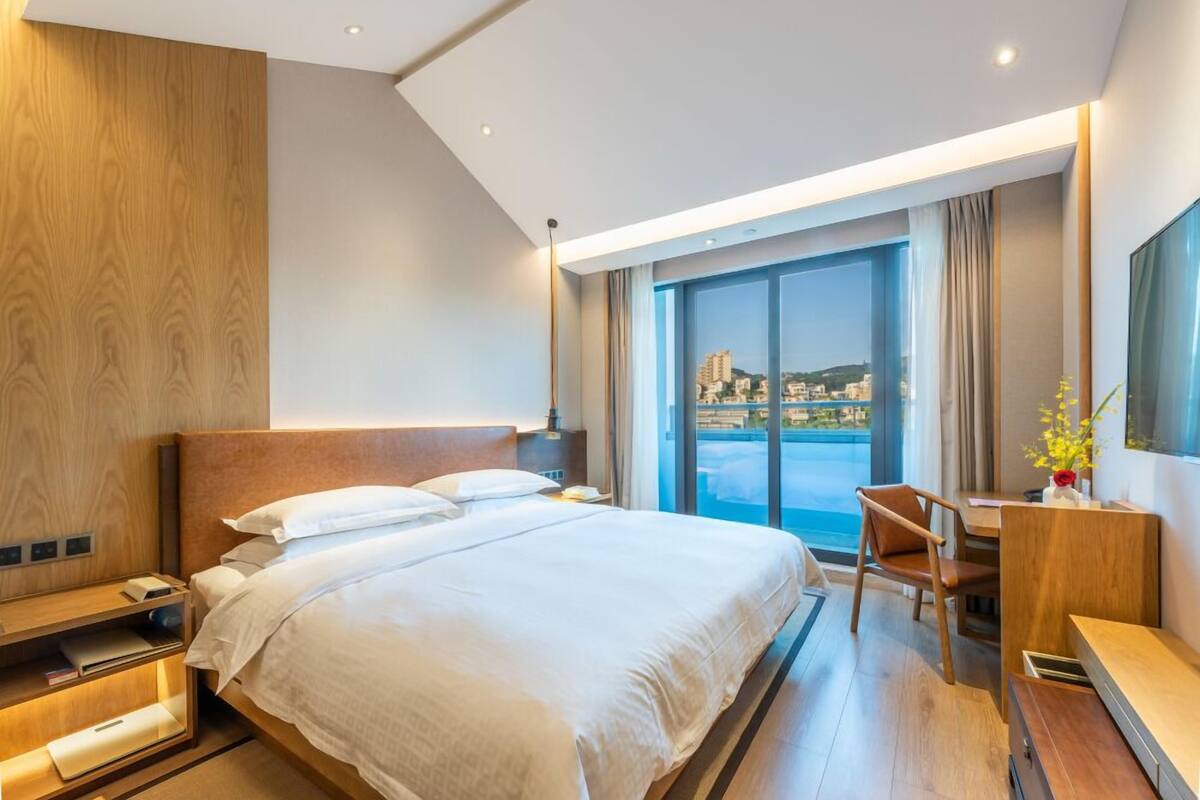 A modern hotel room with a large bed, a desk, and a view of a cityscape through a large window.