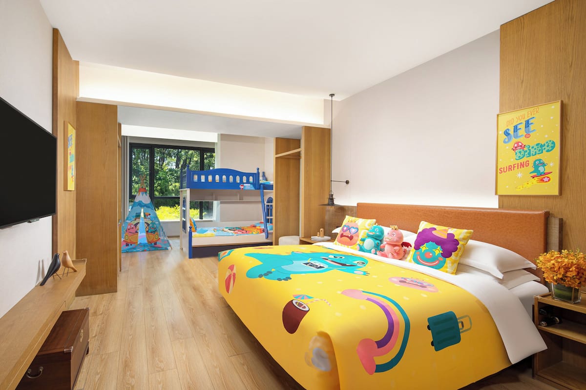 A hotel room with a colorful bedspread, a bunk bed, wooden furniture, and a view of the outdoors.