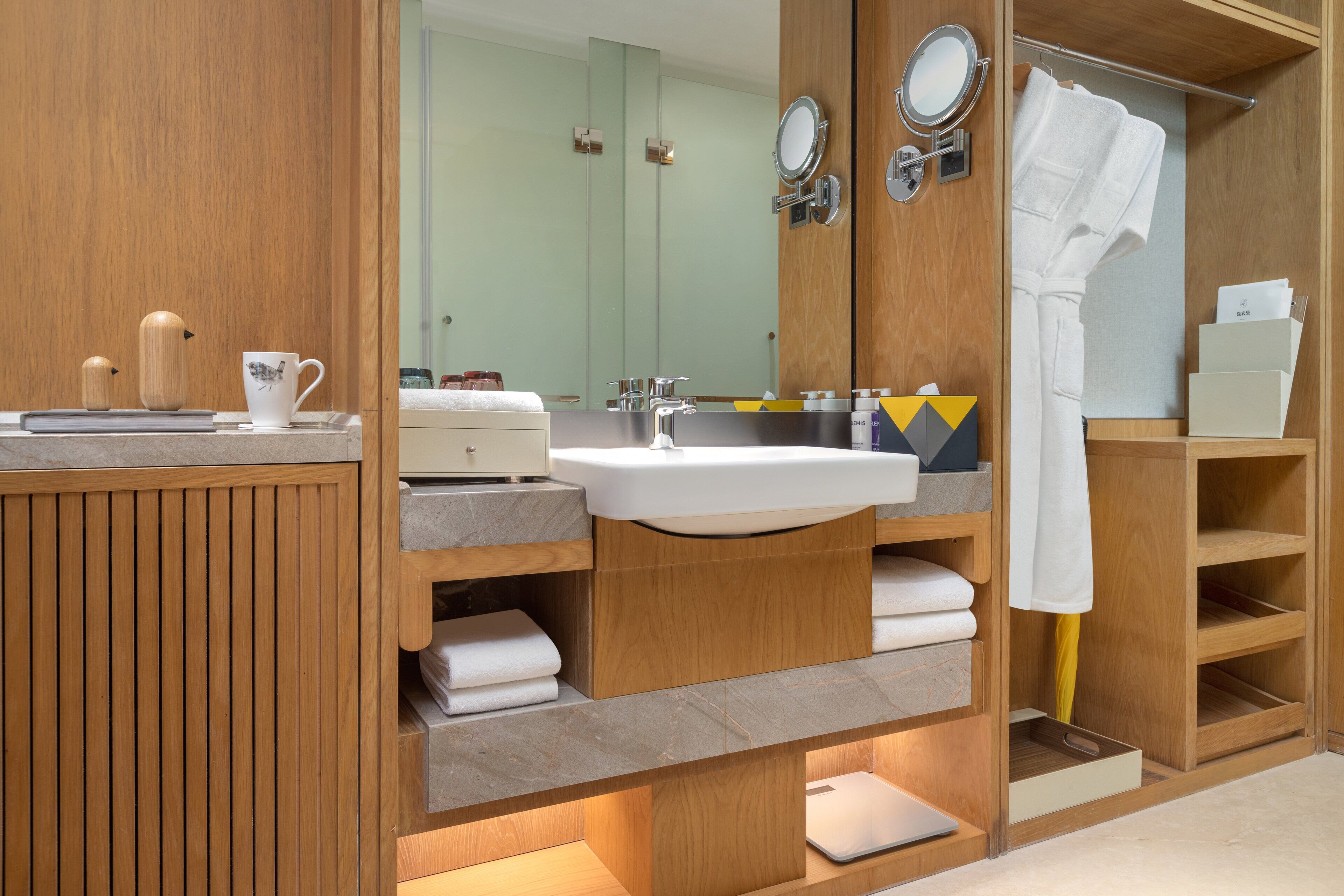 A modern bathroom with a wooden vanity, a sink, and a mirror. There are towels and toiletries on the shelves.