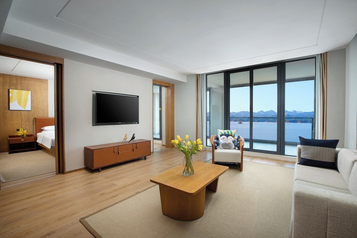 A modern hotel room with a large window, a flat-screen TV, a wooden coffee table, and a sofa.
