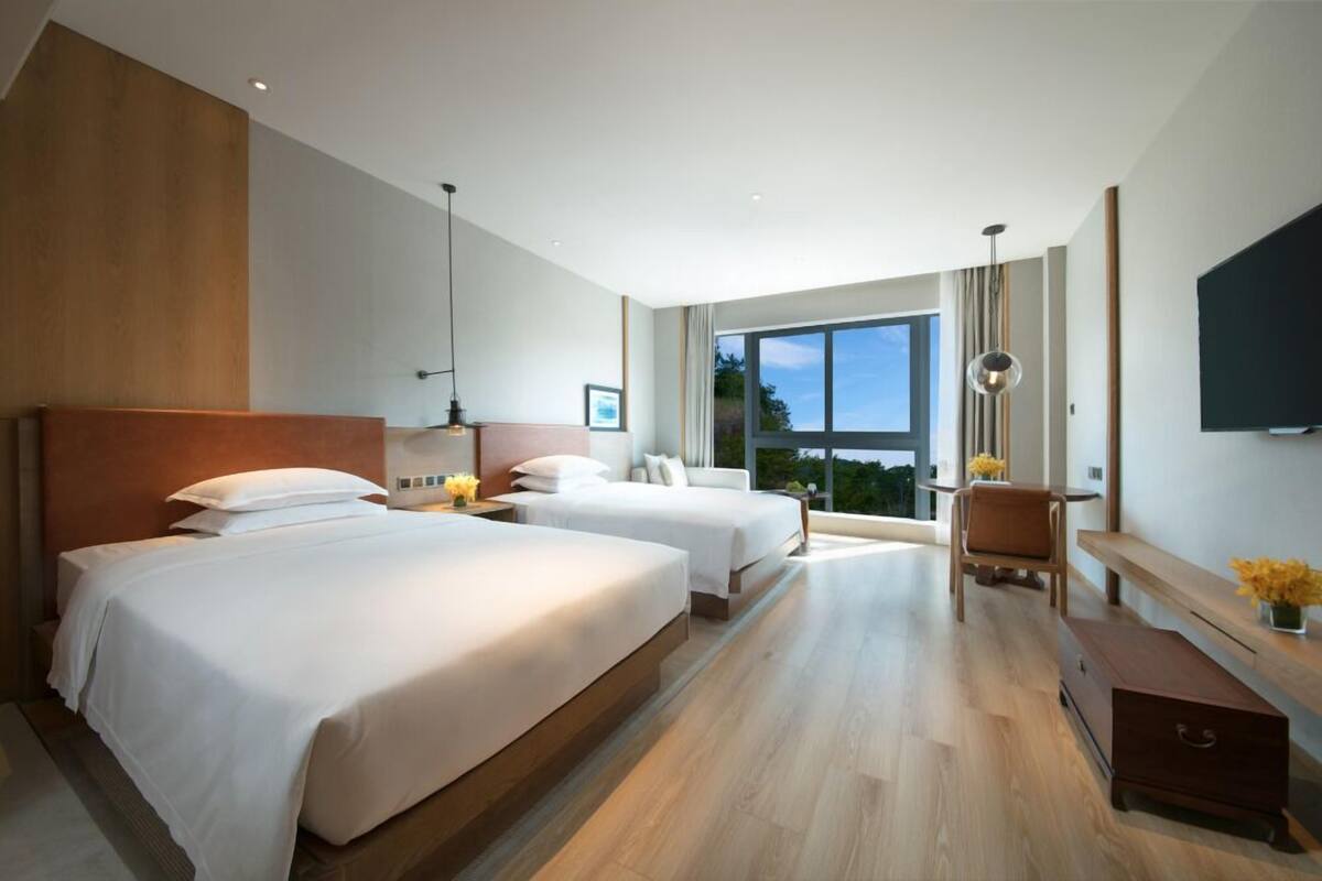 A modern hotel room with two beds, a desk, a chair, and a large window offering a view of the outdoors.