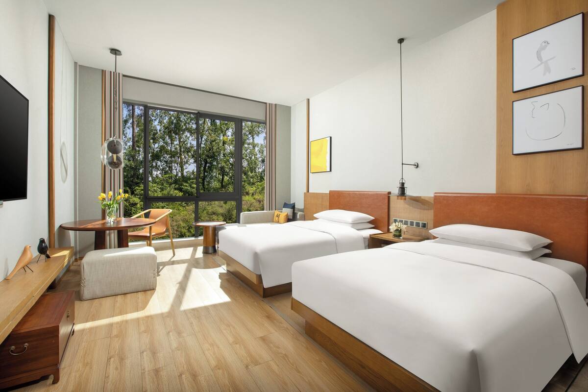 A modern hotel room with two beds, a desk, and a large window offering a view of trees.