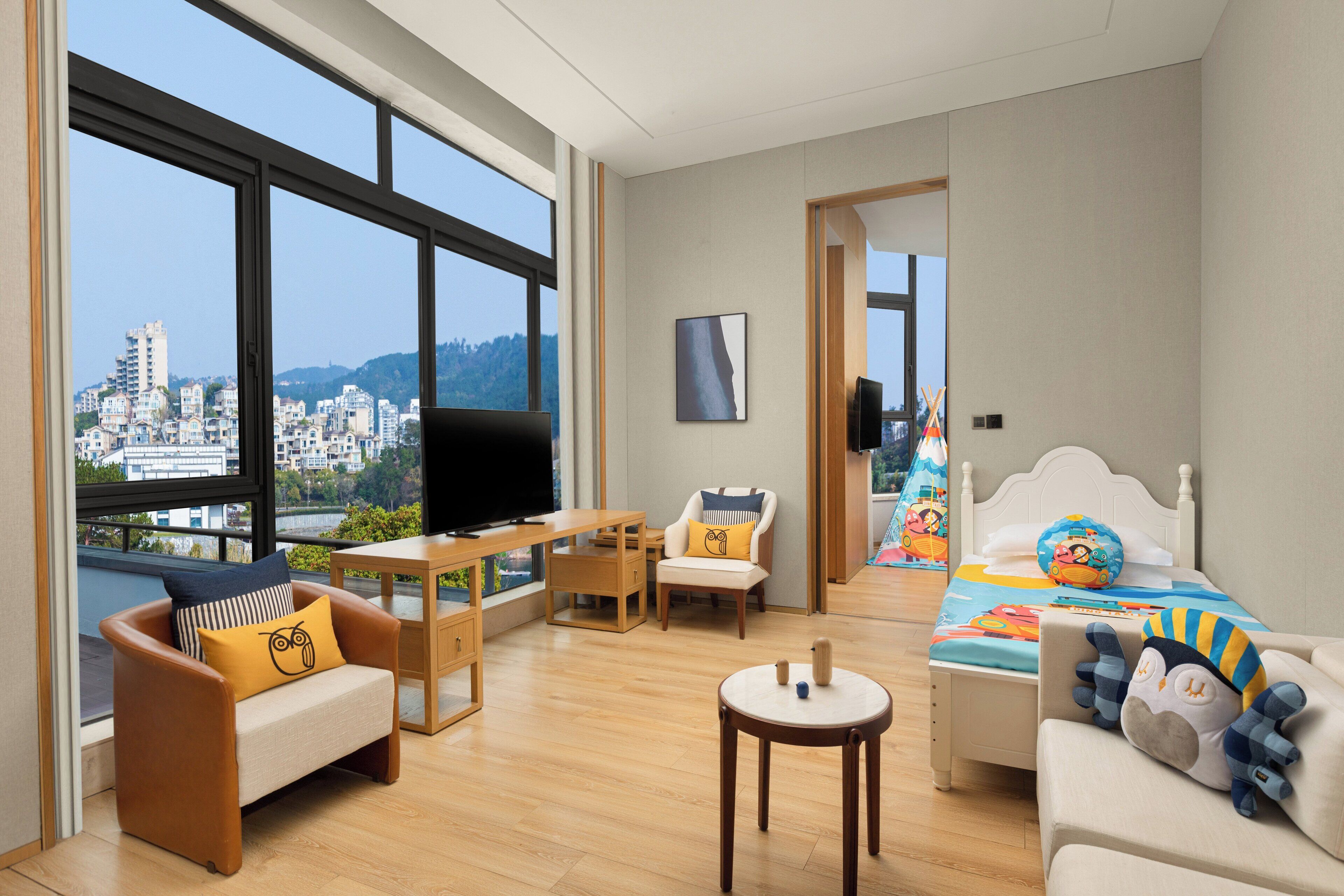 Ett modernt hotellrum med ett stort fönster, en TV, ett skrivbord, en soffa och en säng med ett färgglatt överkast.