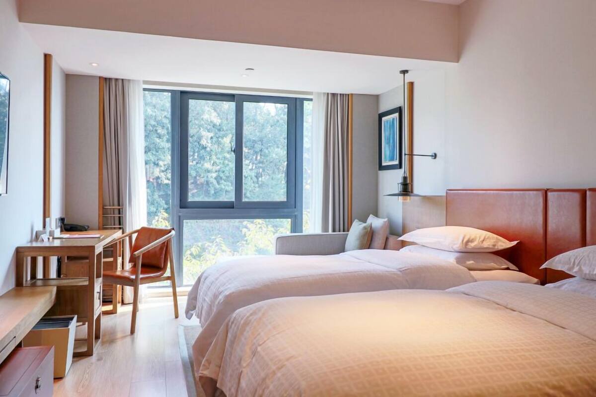 A hotel room with two beds, a desk, and a chair. There is a large window providing a view of trees.