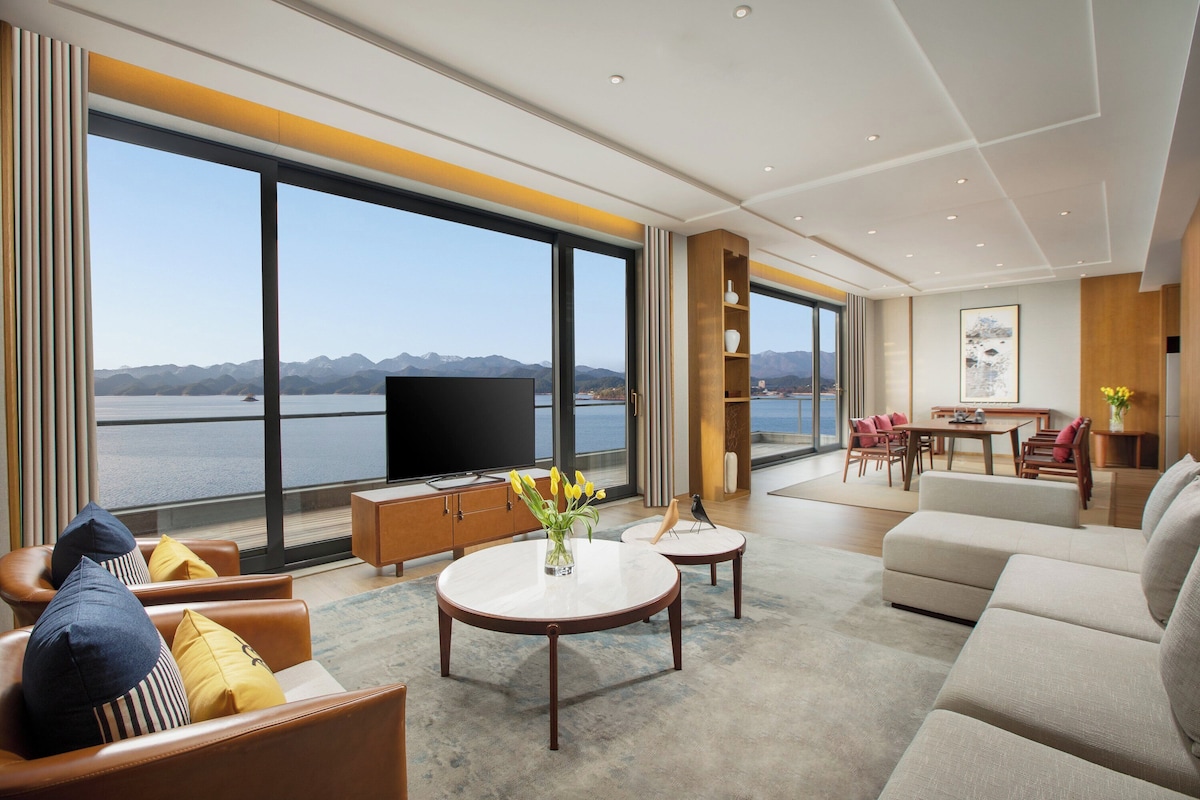 A spacious living room with a large TV, comfortable seating, a dining area, and a view of a lake and mountains.