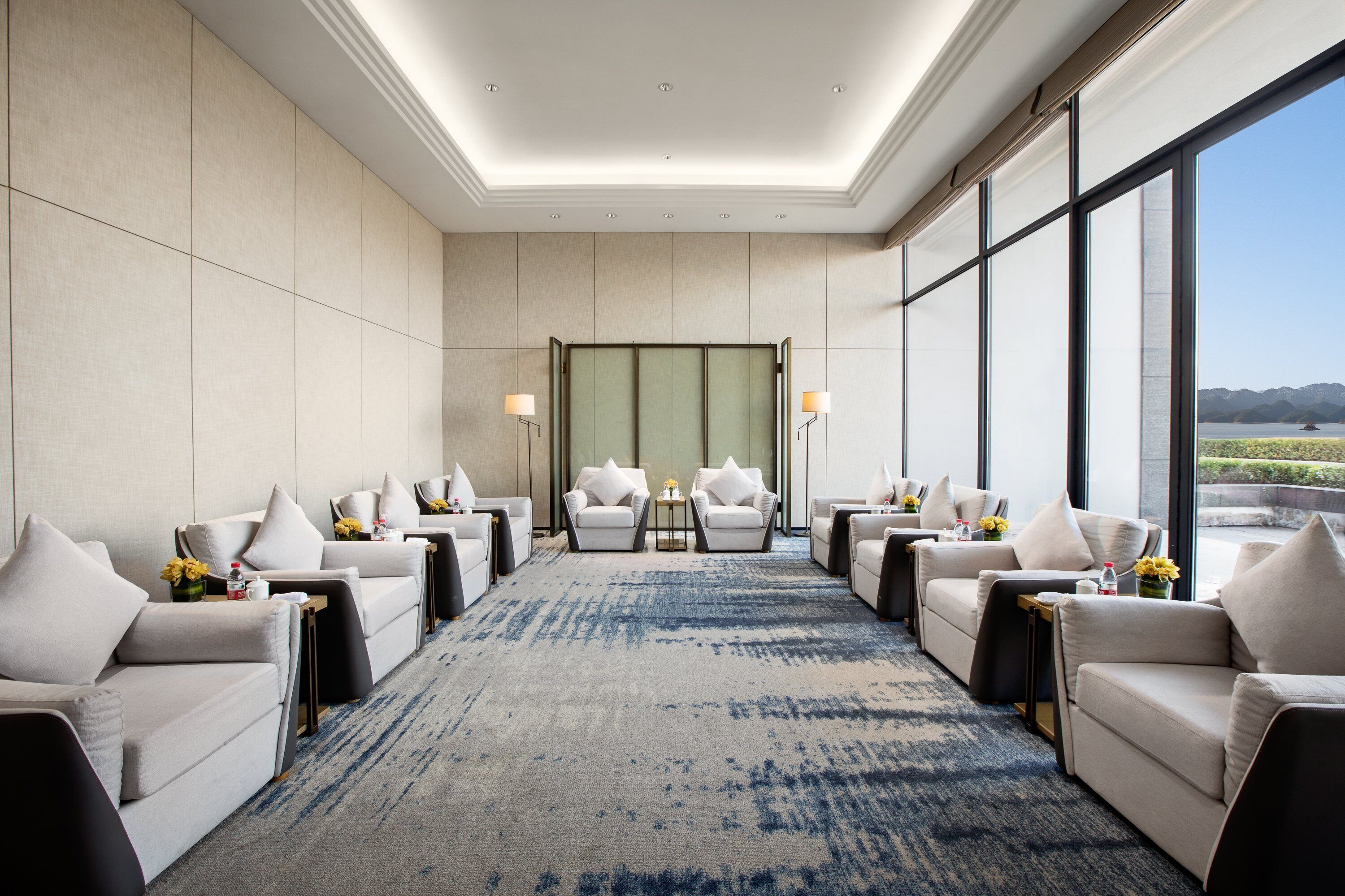 A modern hotel room with a large window, a blue carpet, and multiple seating areas.