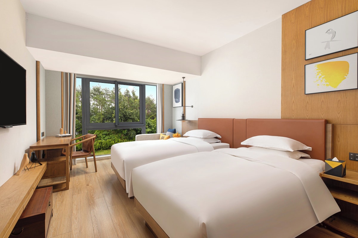 A modern hotel room with two beds, a desk, a chair, and a large window offering a view of greenery.