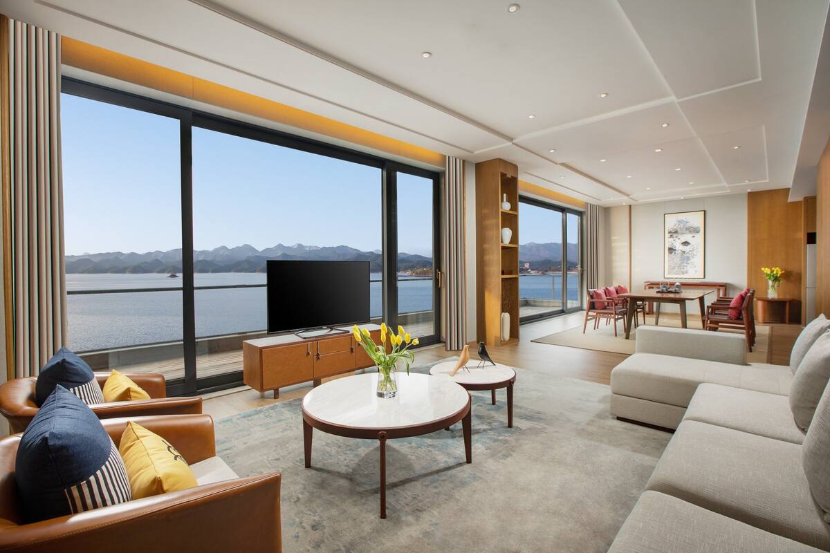 A spacious living room with a large TV, comfortable seating, a dining area, and a view of a lake and mountains.