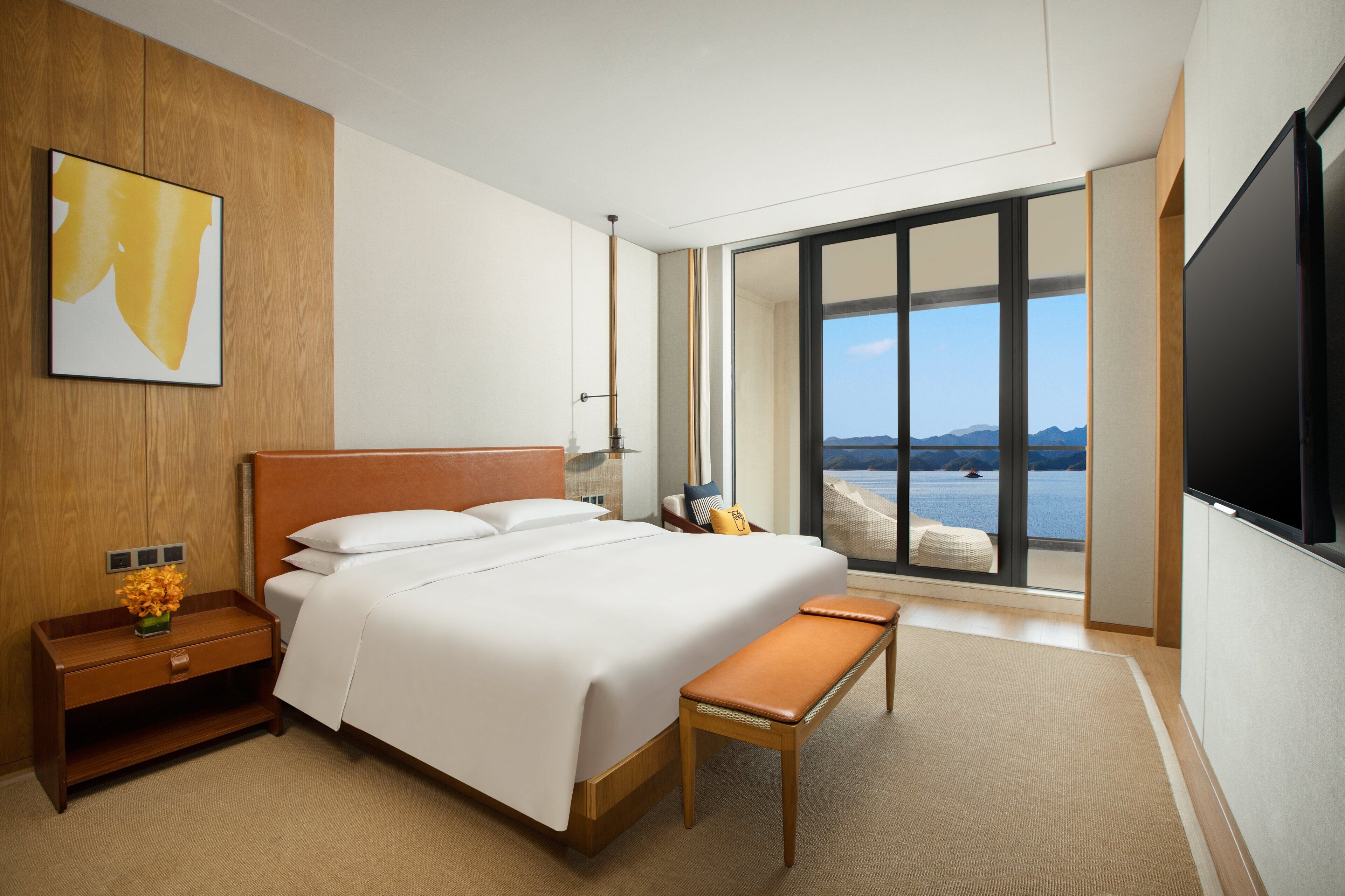 A modern hotel room with a large bed, a wooden nightstand, a bench, and a large window offering a view of the sea.