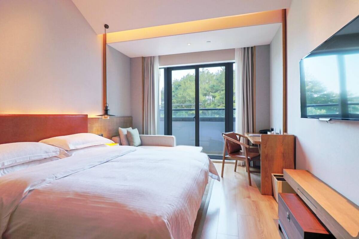 A modern hotel room with a large bed, a desk, and a chair. There is a window with a view of trees and a balcony.