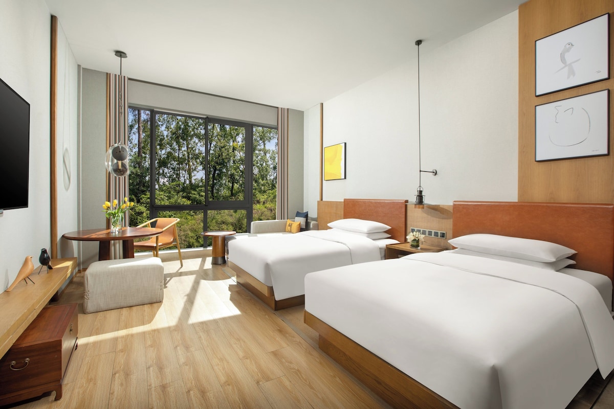 A modern hotel room with two beds, a desk, and a large window offering a view of trees.