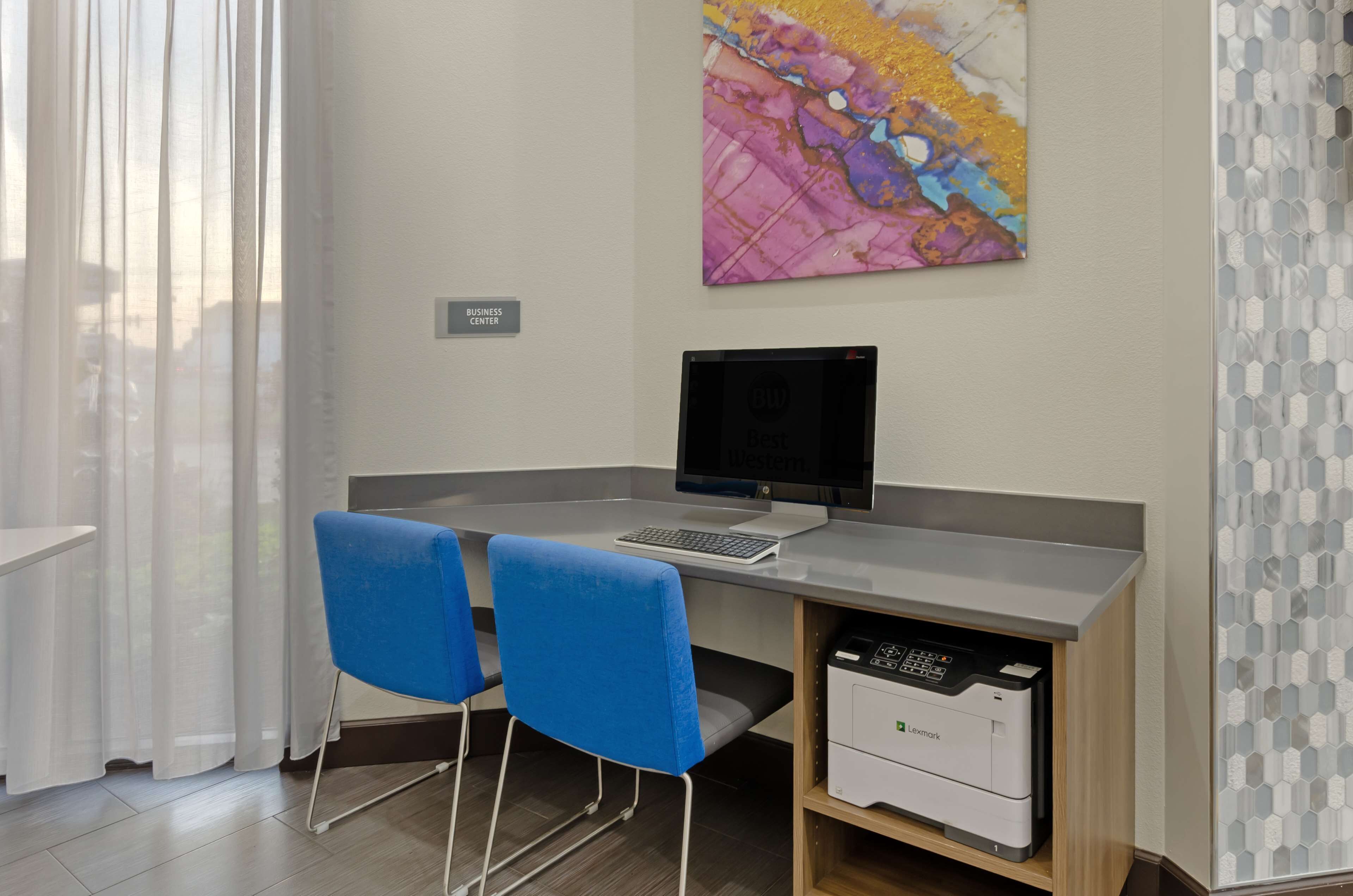 Een business center met een bureau, twee blauwe stoelen, een computer, een printer en een kleurrijk abstract schilderij aan de muur.