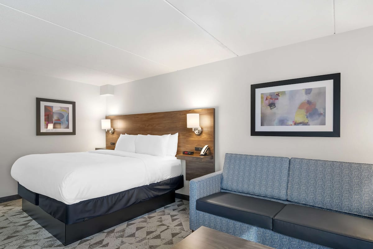 Une chambre d’hôtel avec un grand lit, un canapé bleu, une tête de lit en bois, deux lampes fixées au mur et deux œuvres d’art encadrées.