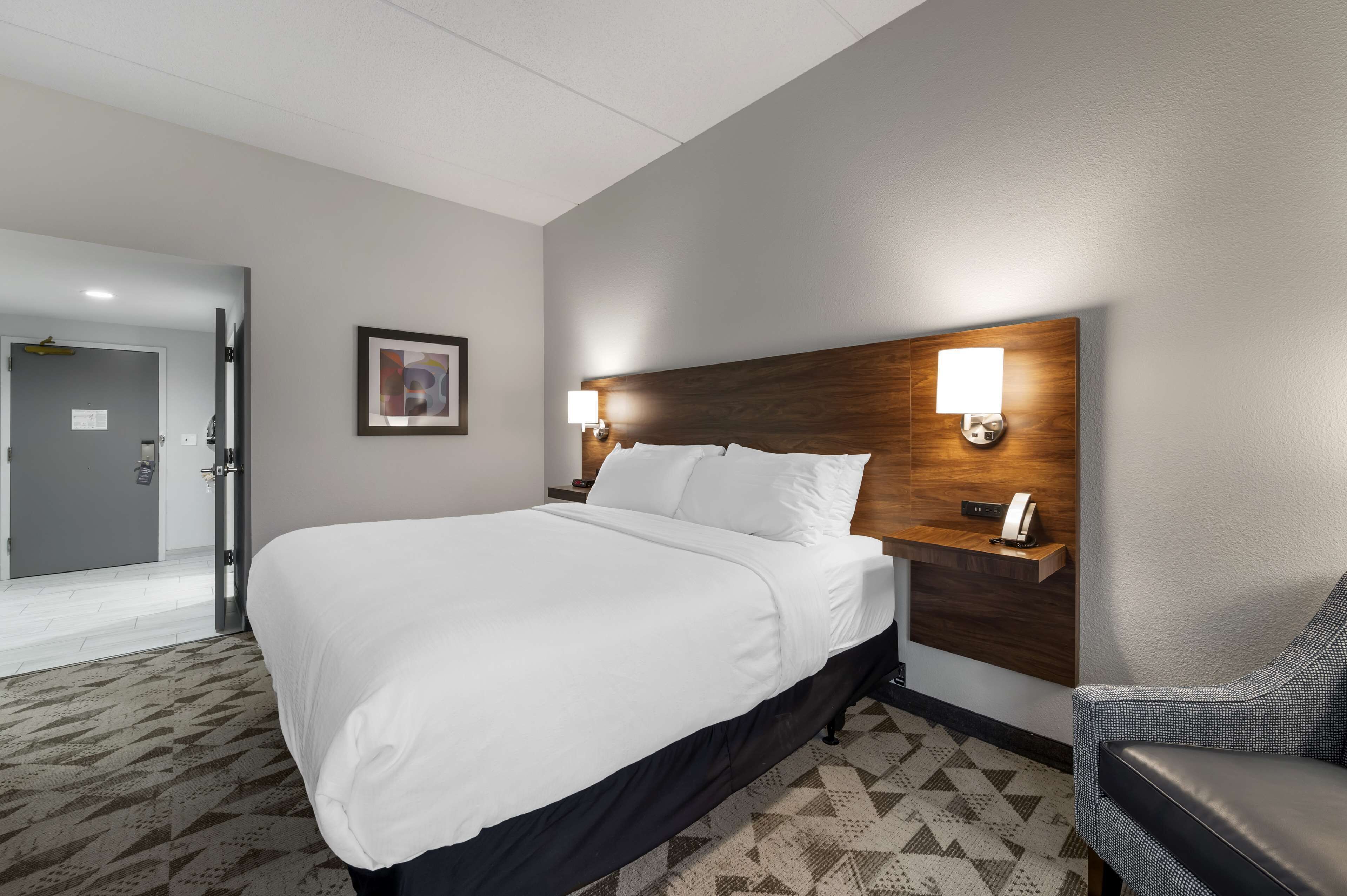 A hotel room with a large bed, a wooden headboard, two bedside lamps, a desk with a phone, and a patterned carpet.