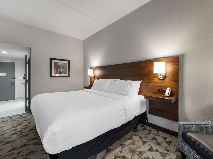 A hotel room with a large bed, a wooden headboard, two bedside lamps, a desk with a phone, and a patterned carpet.