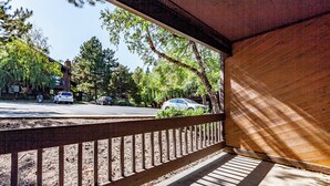 A covered outdoor area with a wooden railing, a paved walkway, and a view of parked cars and trees.