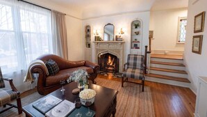Ein gemütliches Wohnzimmer mit Kamin, einer Ledersofa, einem Karo-Sessel und einem Esstisch mit Weingläsern und einer Speisekarte.