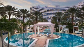 Una piscina grande y curvada con sillas de descanso y un pabellón, rodeada de palmeras y un edificio de hotel al fondo.