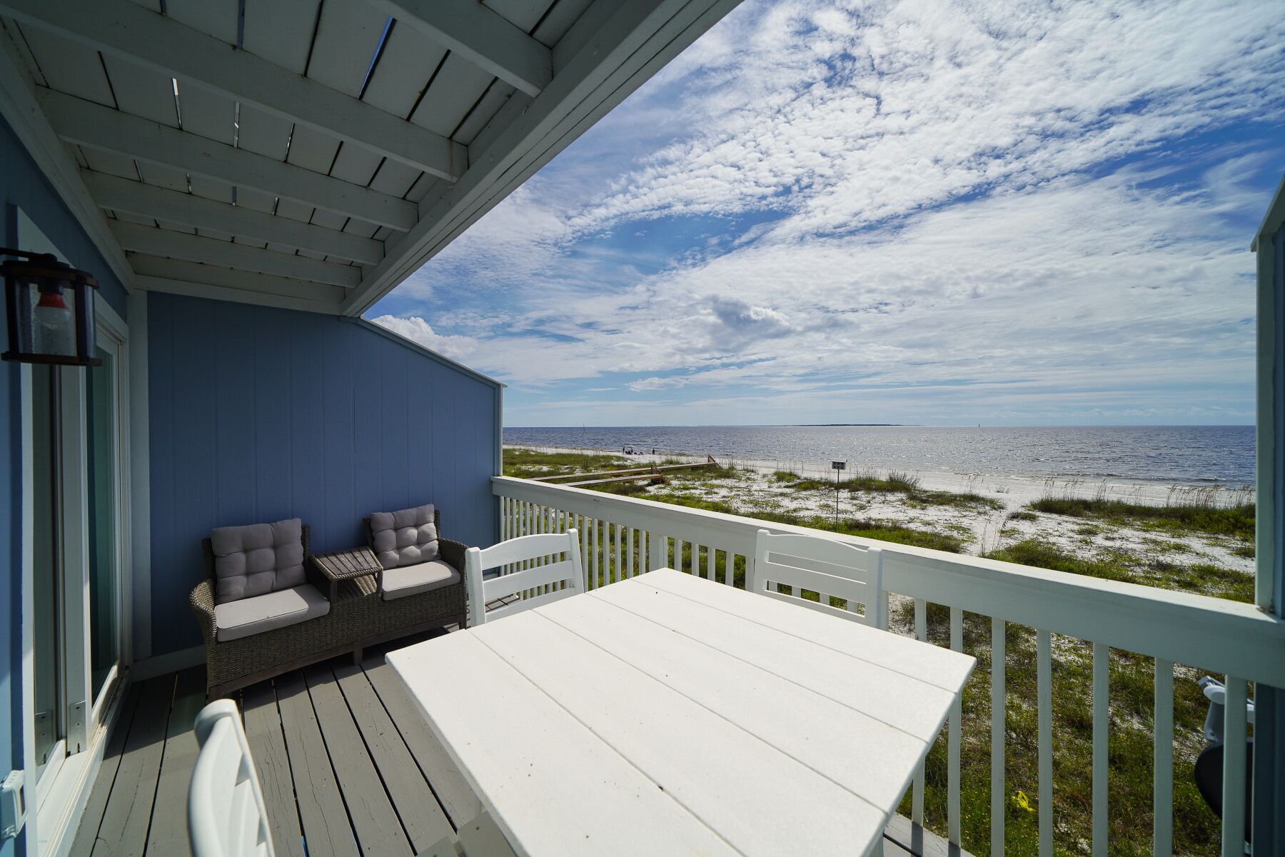 Une terrasse couverte avec une table blanche et deux chaises rembourrées, donnant sur une plage de sable avec une végétation clairsemée.