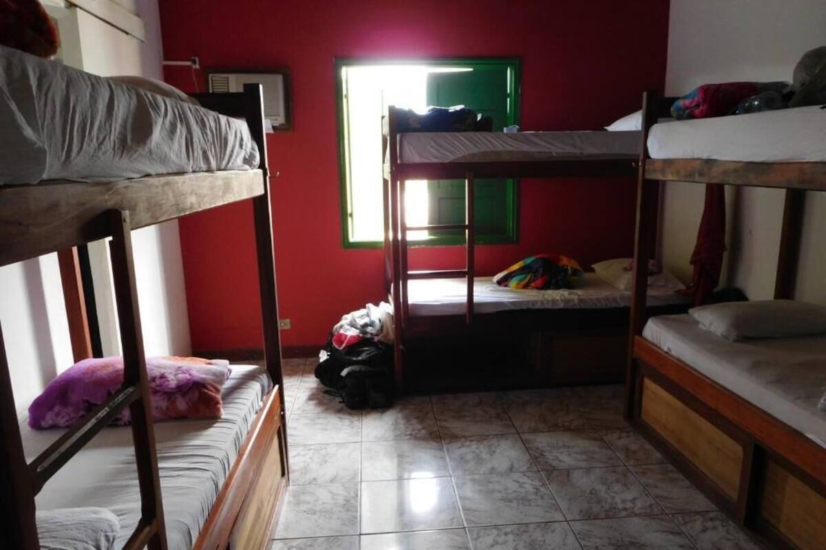 A dormitory room with bunk beds, a window with a green frame, and a tiled floor.
