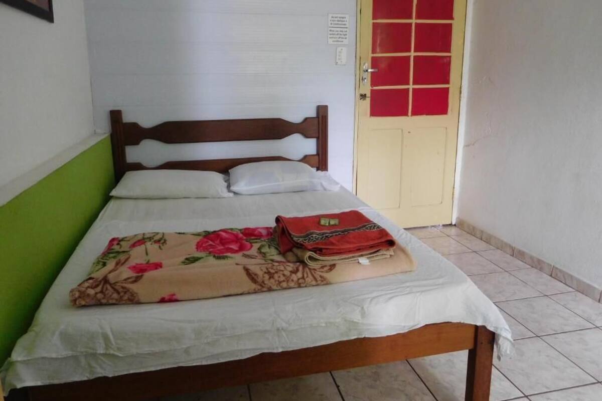 A single bed with a floral bedspread, a folded red blanket, and a white pillow. A red-framed door is visible in the background.