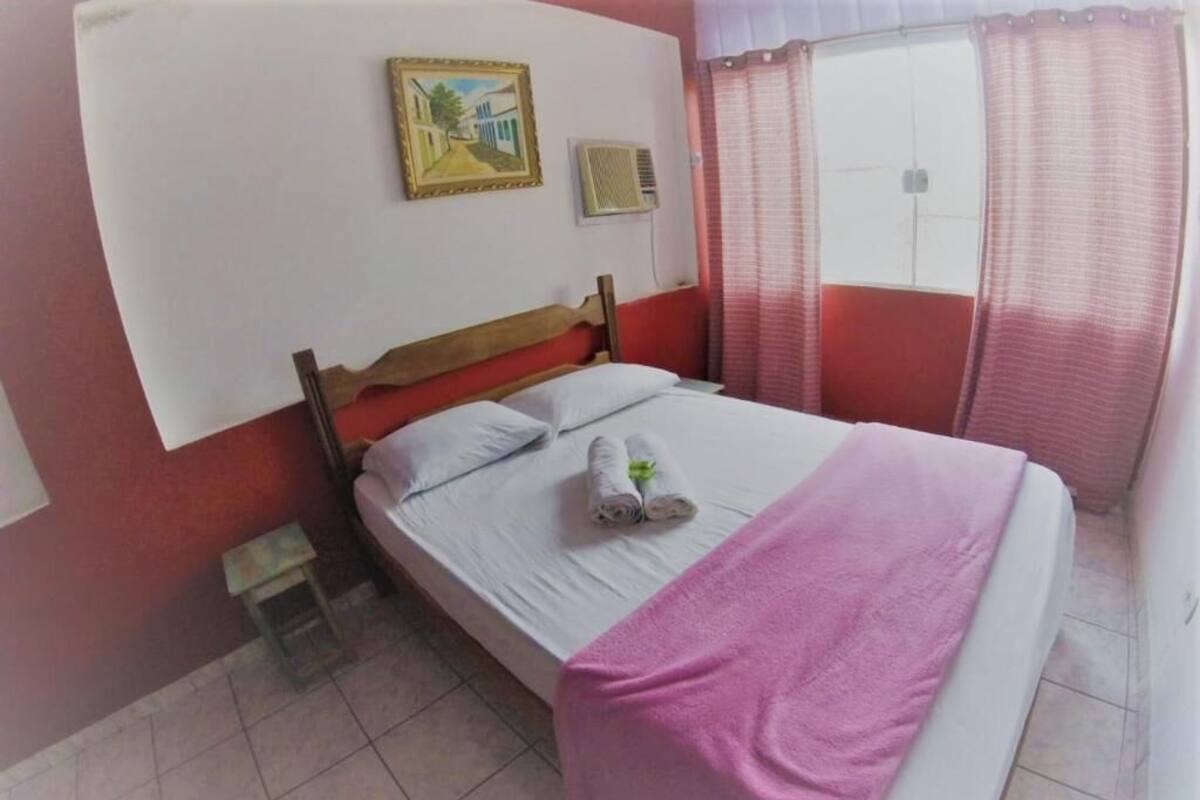 A bedroom with a bed, a small bedside table, a wall-mounted air conditioner, a window with curtains, and a framed picture on the wall.