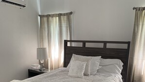 A neatly made bed with a dark wooden headboard, a white throw blanket, and a matching pillow set. There is a bedside table with a lamp and a folded towel. The room has light-colored walls and a carpeted floor. - Sapphire Villa Near the Sea-Entire Home (Nassau)