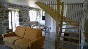 Una habitación con pared de piedra, una escalera de madera, un sofá amarillo y una mesa de comedor con sillas.
