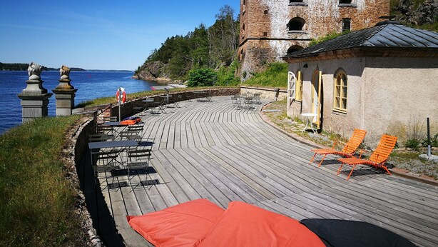 A wooden deck with outdoor seating, a brick building, and a view of the water. - Fredriksborg Hotel (Värmdö)
