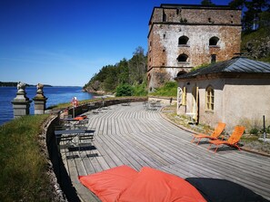 A wooden deck with outdoor seating, a brick building, and a view of the water.