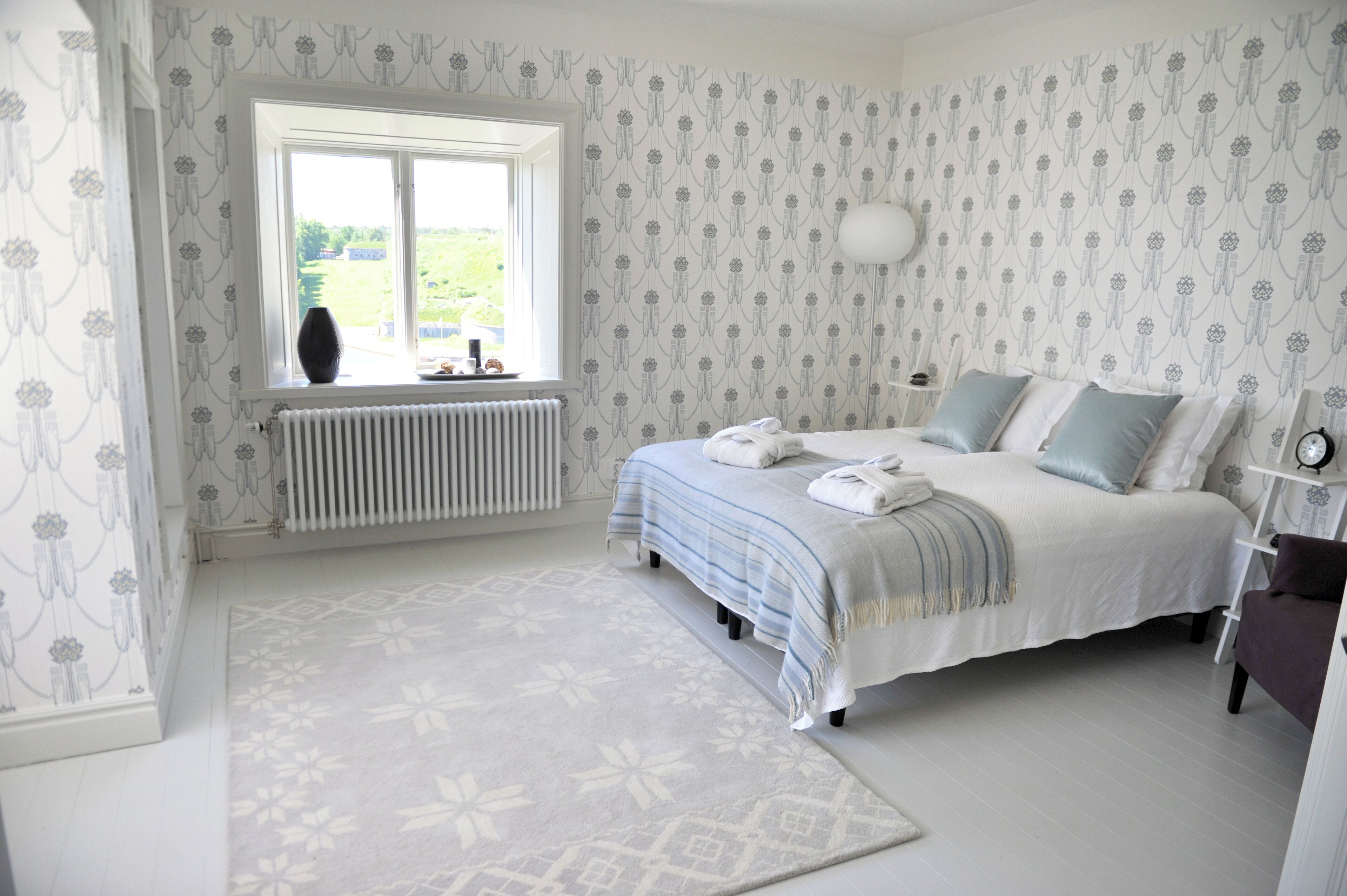 A bedroom with a large window, a radiator, a bed with pillows, a bedside table, and a chair.