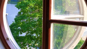 A round window with a wooden frame and a clear glass pane, offering a view of a lake surrounded by lush green trees. - Fredriksborg Hotel (Värmdö)