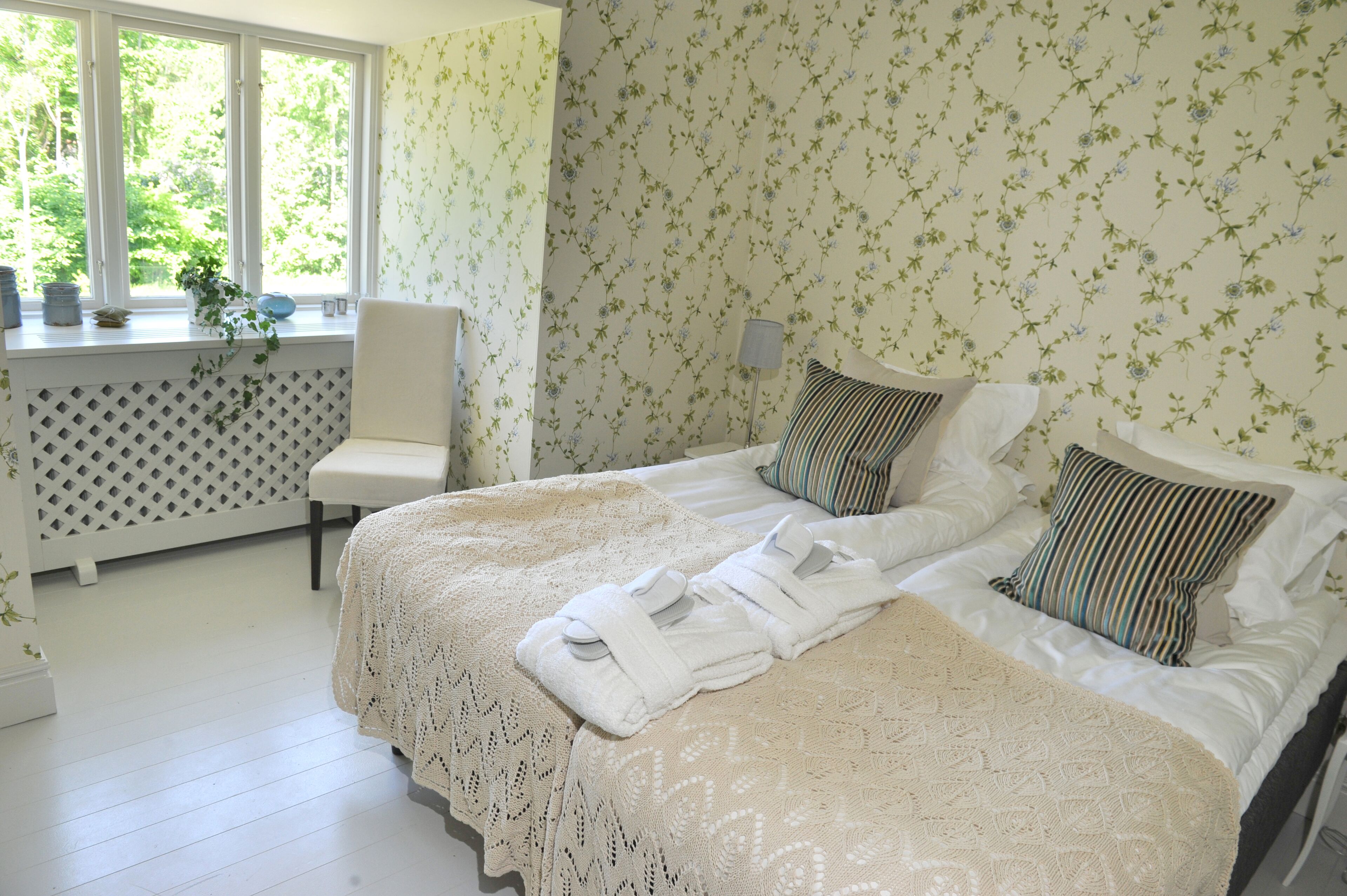 Un dormitorio con una cama, dos almohadas, una mesita de noche, una silla y una ventana con plantas.