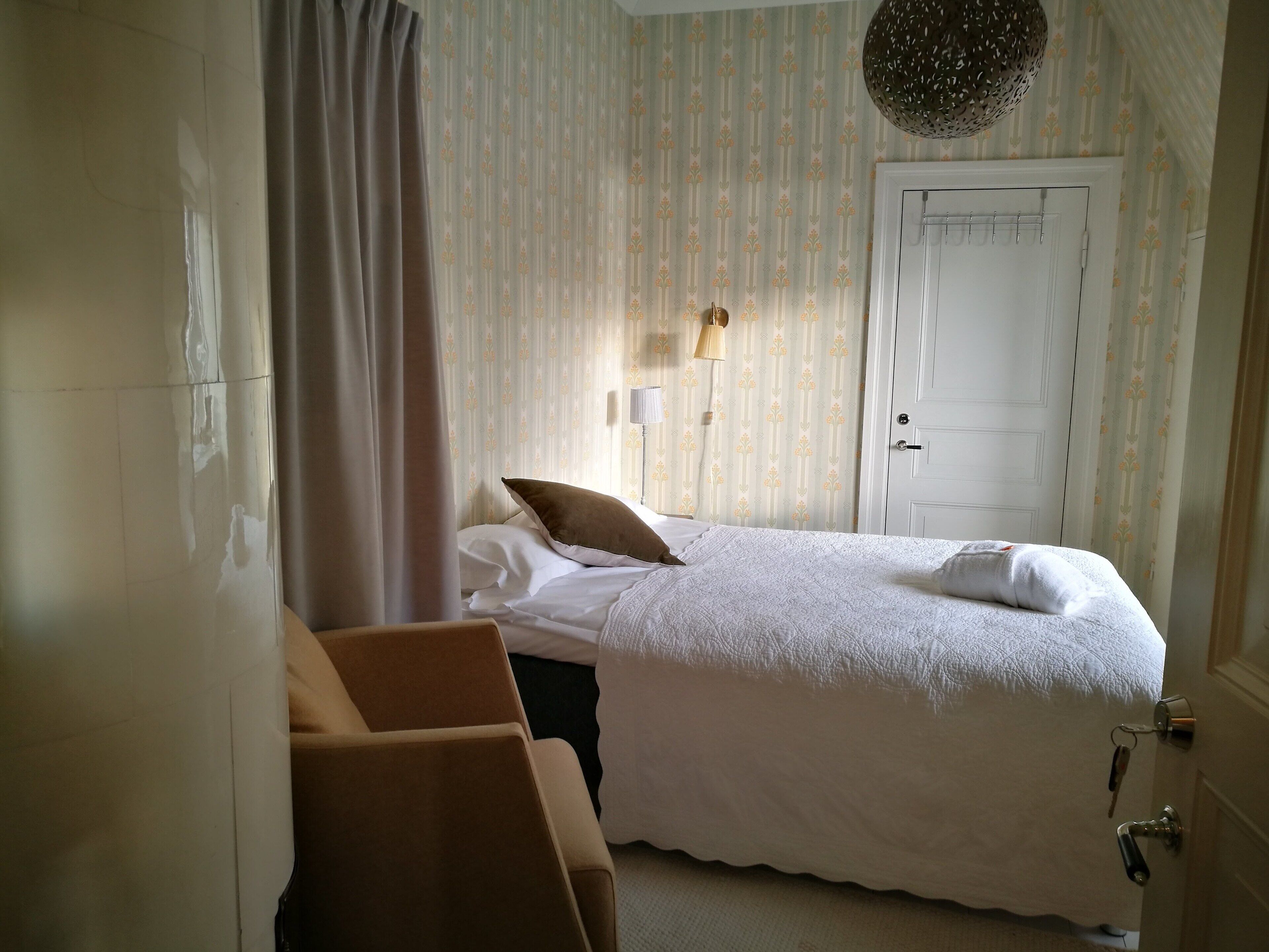 Ein ordentlich bezogenes Bett mit weißer Decke und Kissen, eine Nachttischlampe und eine Tür mit Schlüssel im Schloss.