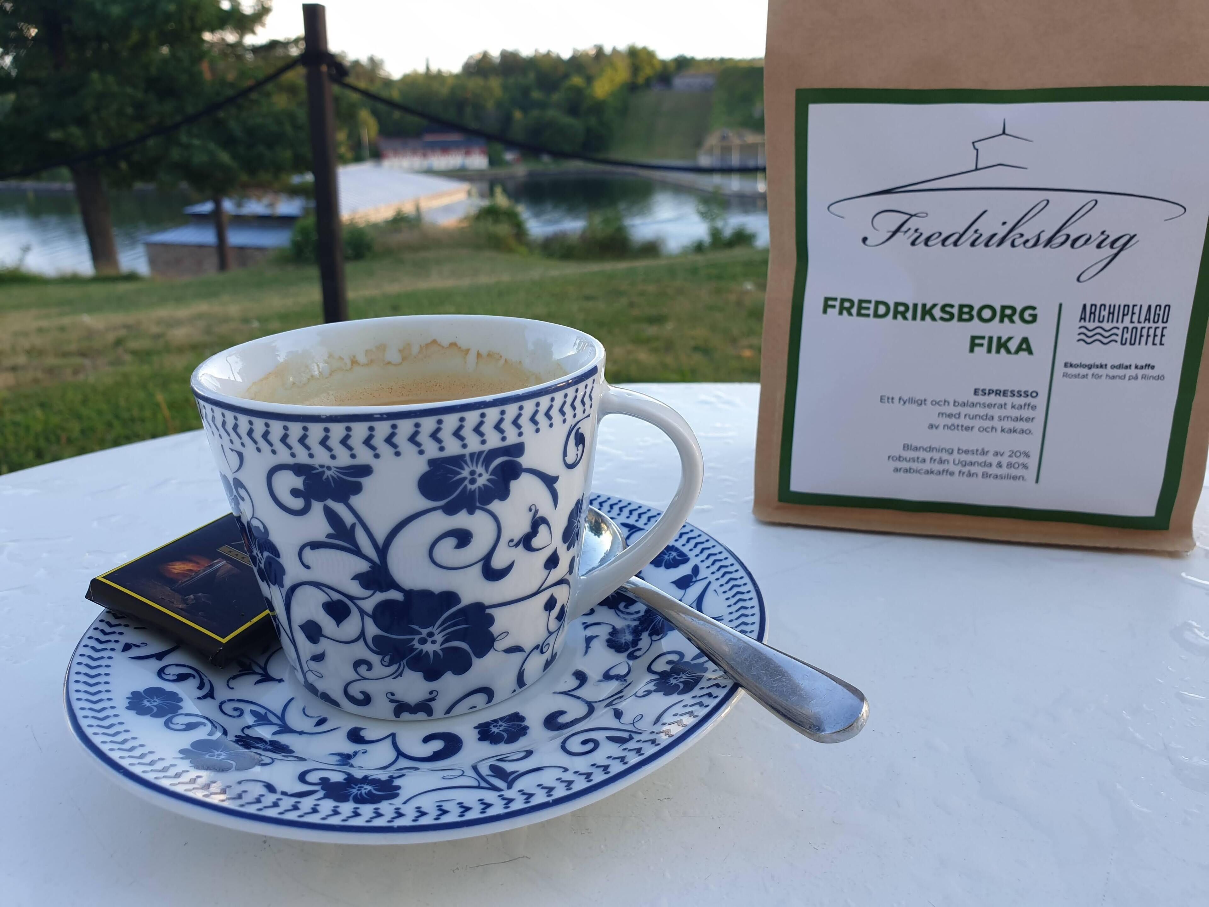 A cup of coffee on a saucer with a spoon, a coffee bag, and a menu on a table with a view of a lake and trees in the background.