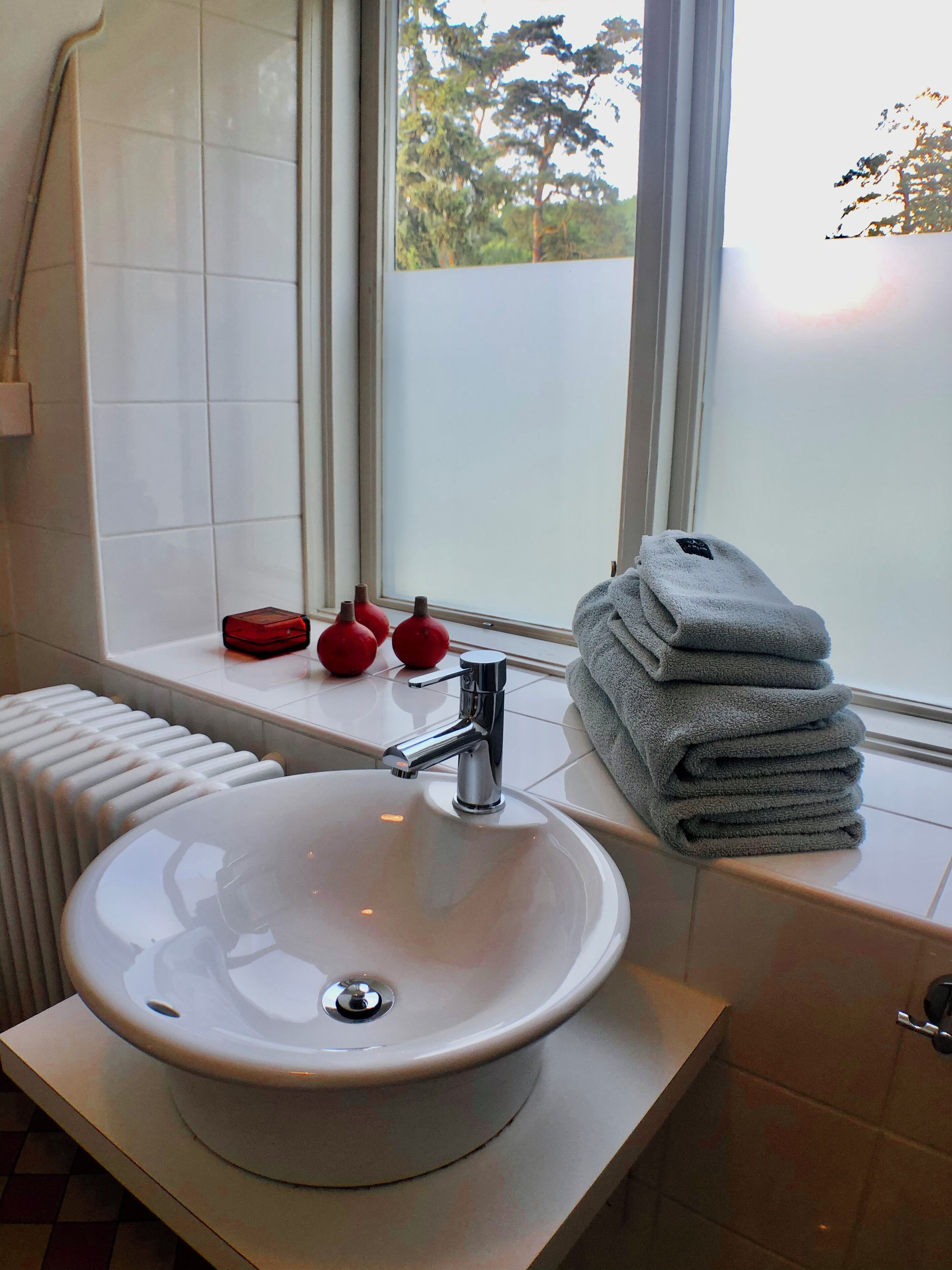 Ein Badezimmer mit einem weißen Waschbecken, einem Spiegel, einem Fenster mit Blick auf Bäume und einem Stapel Handtüchern am Fensterbrett.